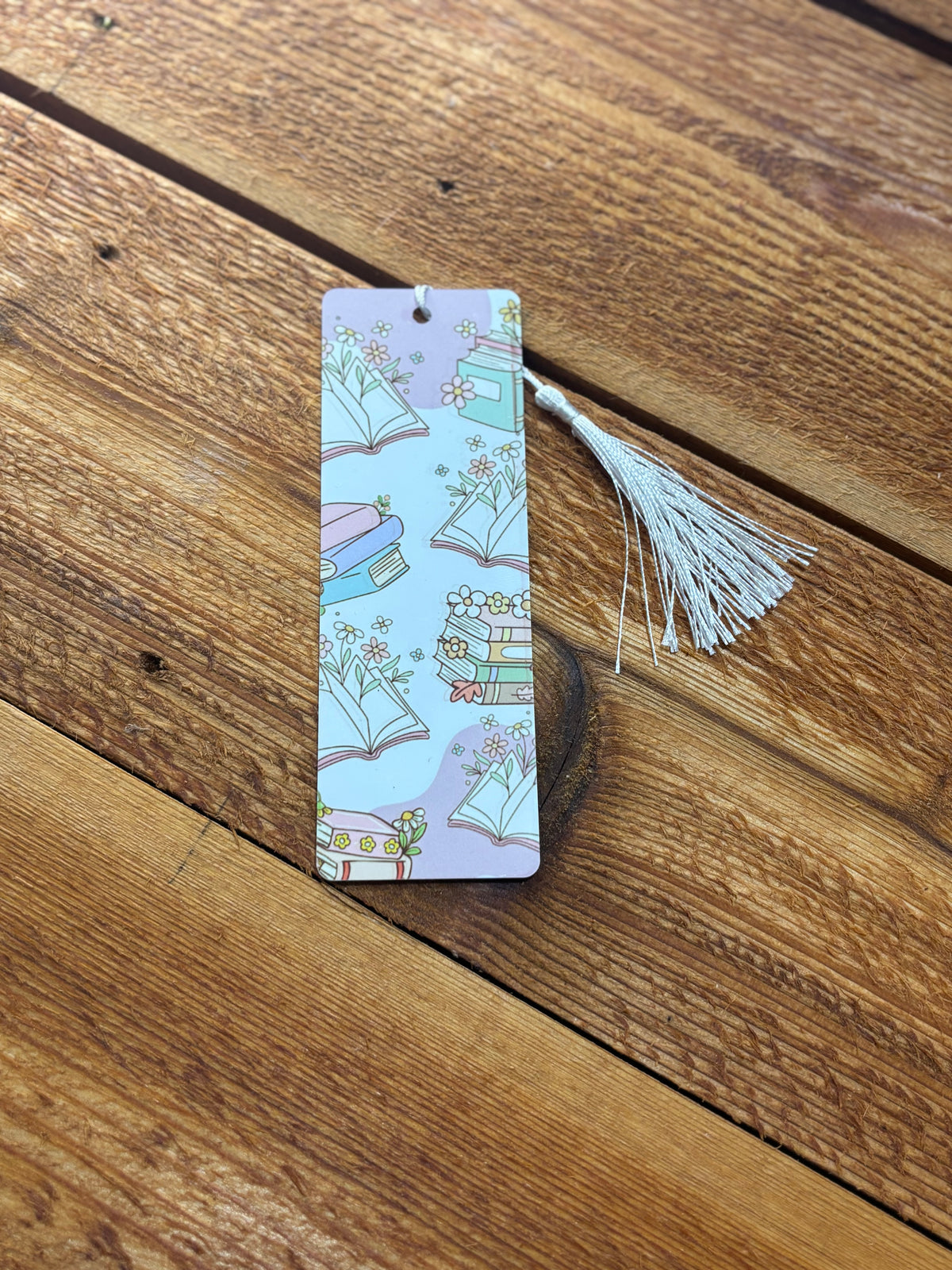 Pastel bookmark with open books, book stacks, flowers, and a white tassel on a wooden surface.
