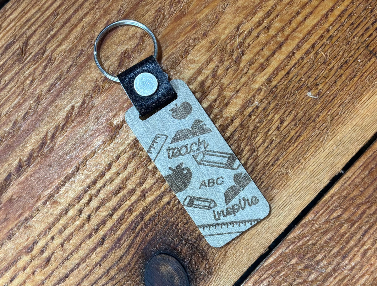 Silver-tone teacher keychain with teach, ABC, inspire, apples, books, pencils, and rulers on a rectangular tag.