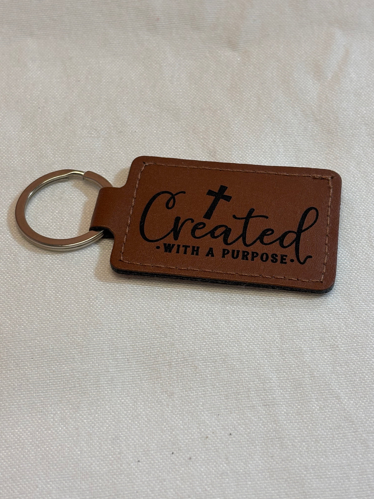 Brown rectangular keychain with stitched edges, engraved with a cross and “Created With a Purpose.”