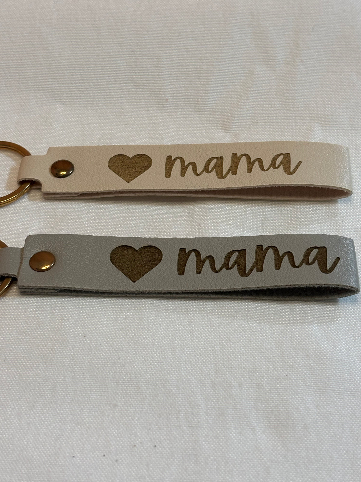Cream and gray wristlet keychains engraved with a heart and “mama” script, finished with gold-tone hardware.