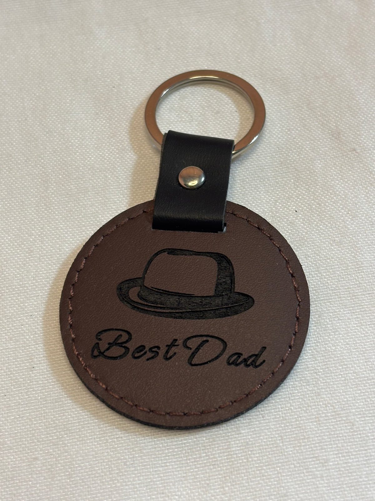 Round brown keychain engraved with a hat and “Best Dad” script, with stitched edges and a black attachment strap.