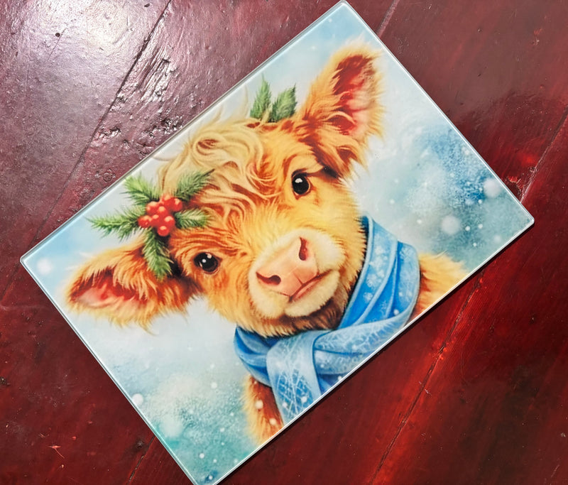 Decorative cutting board with a cow wearing a blue scarf and holly decorations on a wooden surface.