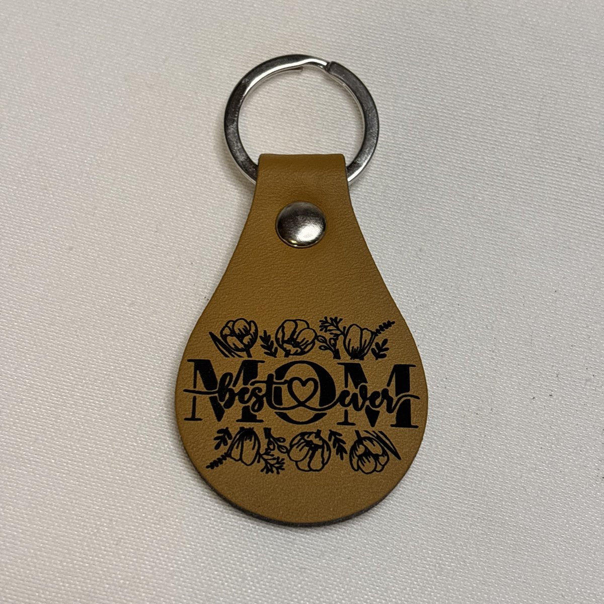 Tan teardrop keychain laser engraved with “Best Mom Ever” lettering, a heart and floral accents.