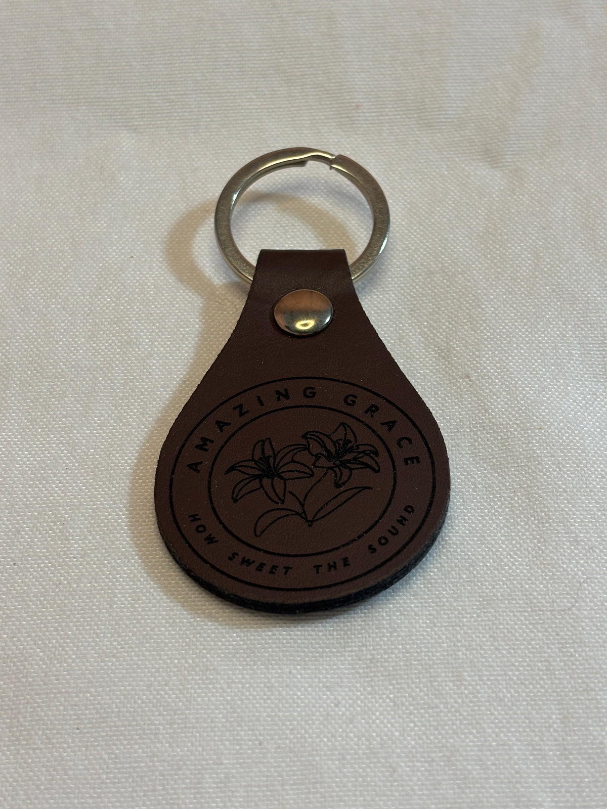Brown teardrop keychain engraved with lilies and “Amazing Grace, How Sweet the Sound” inside a circular border.
