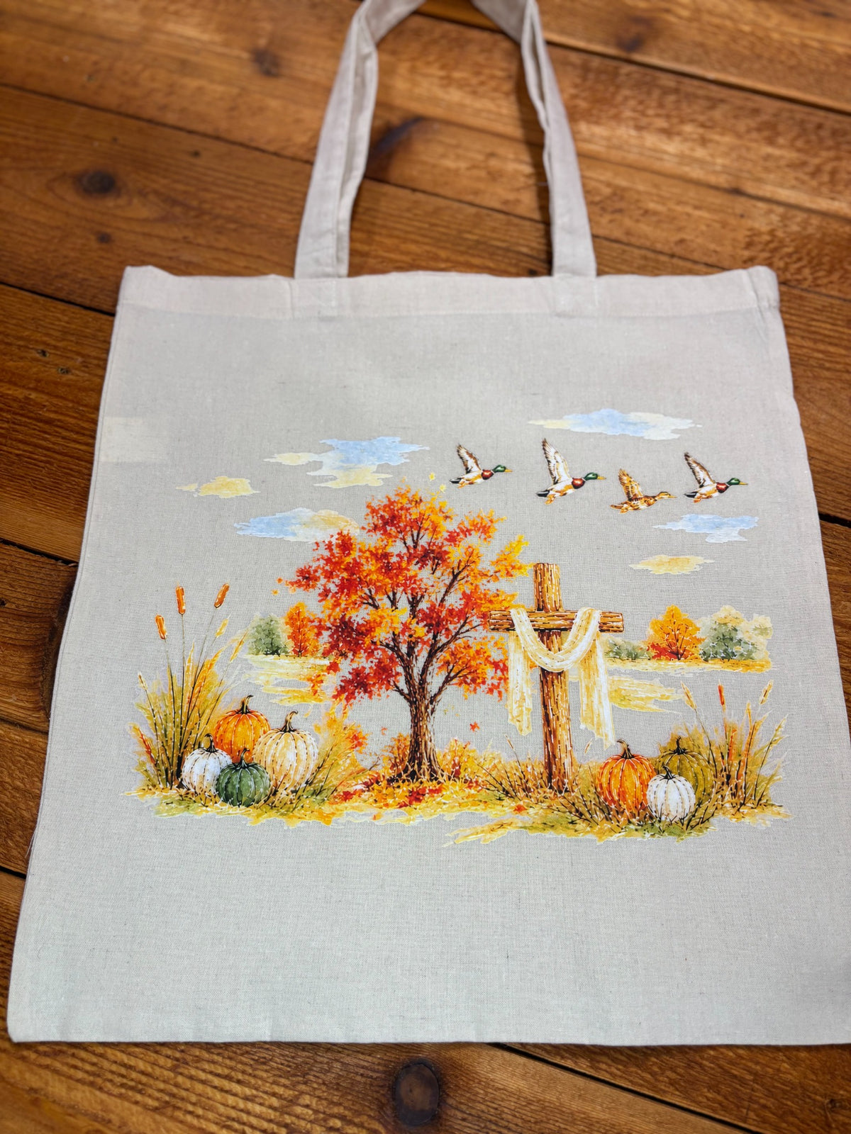 Natural canvas tote bag with watercolor autumn scene featuring a wooden cross, red maple tree, pumpkins, and flying mallard ducks