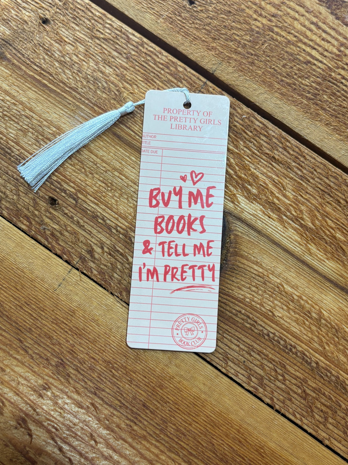 Pink and cream library card bookmark with white tassel reading “Buy me books & tell me I’m pretty” on a wood background