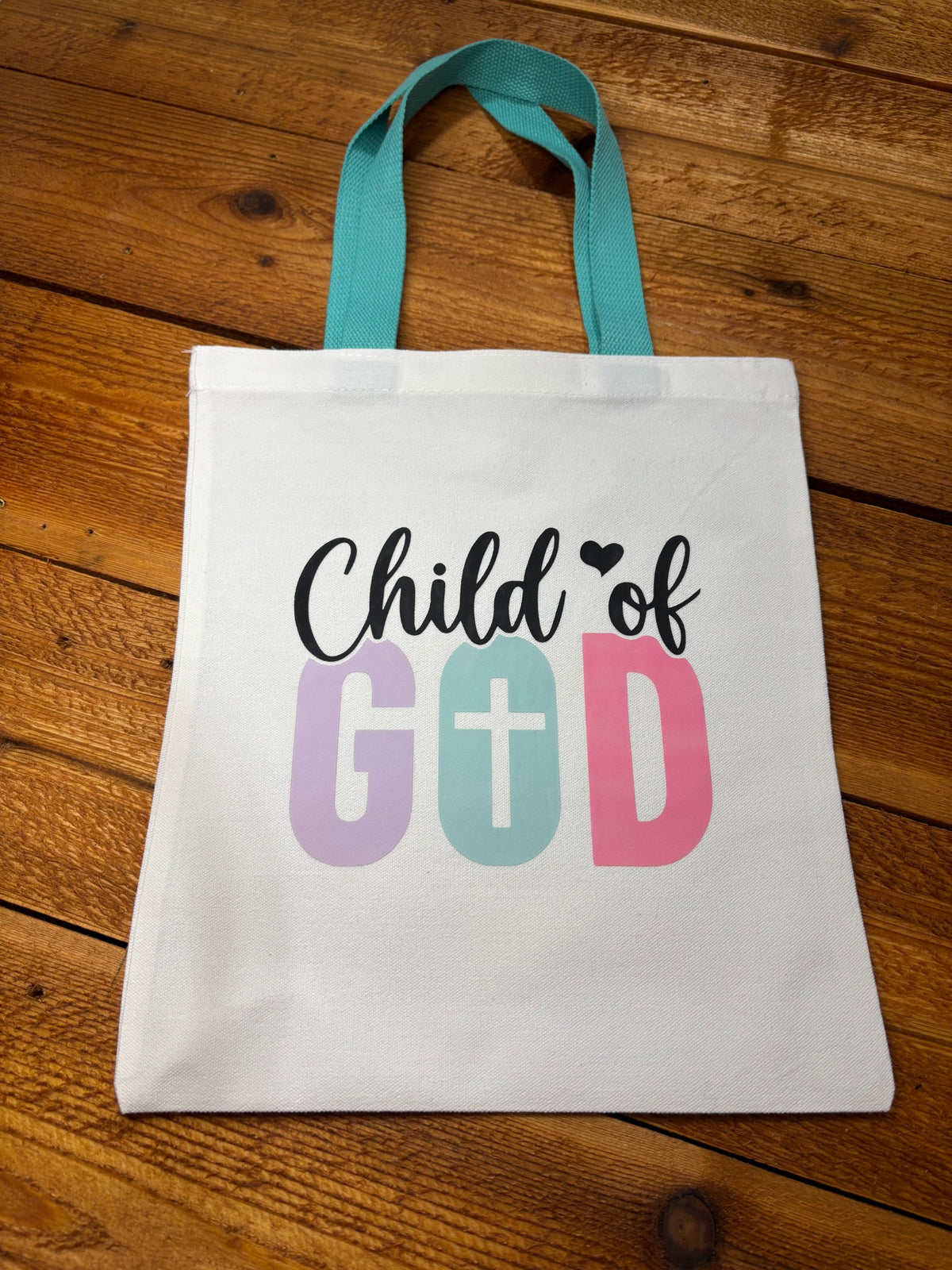 White canvas kids tote bag with teal handles and “Child of God” lettering in purple, mint, and pink with a cross in the O