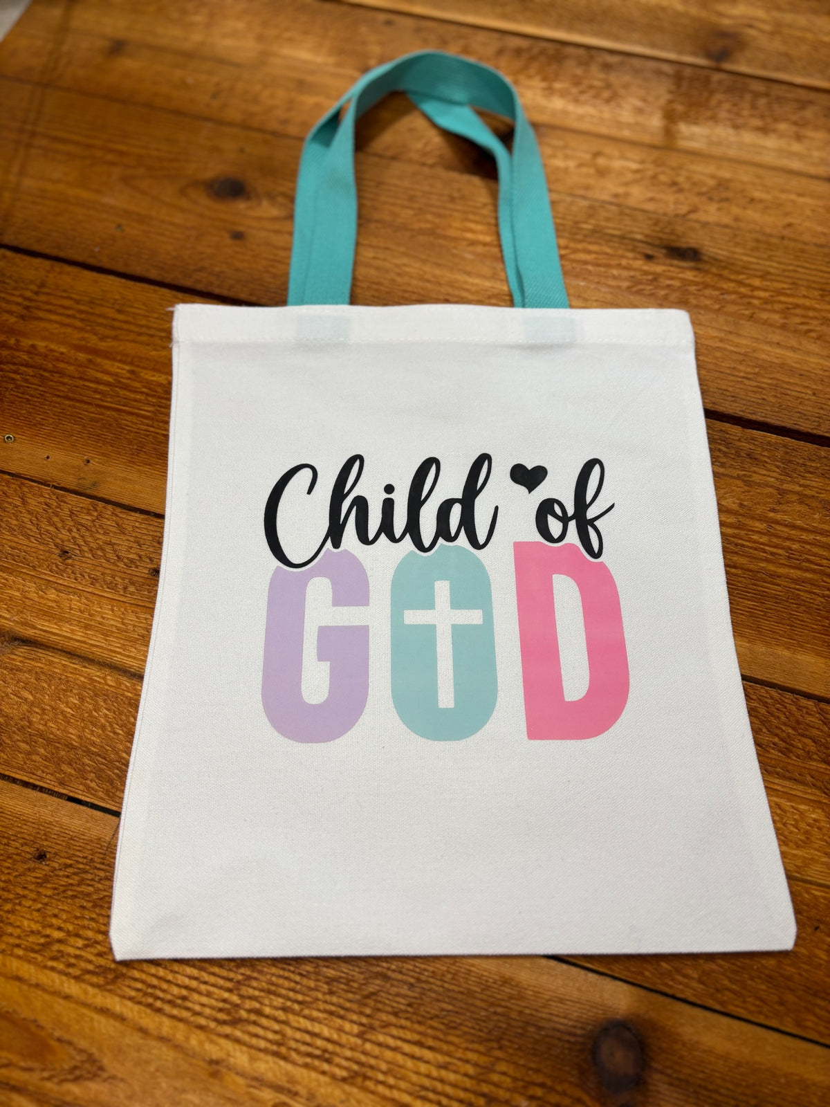 Child of God Christian tote bag for Sunday school with pastel lettering, cross design, and teal handles on a wood surface
