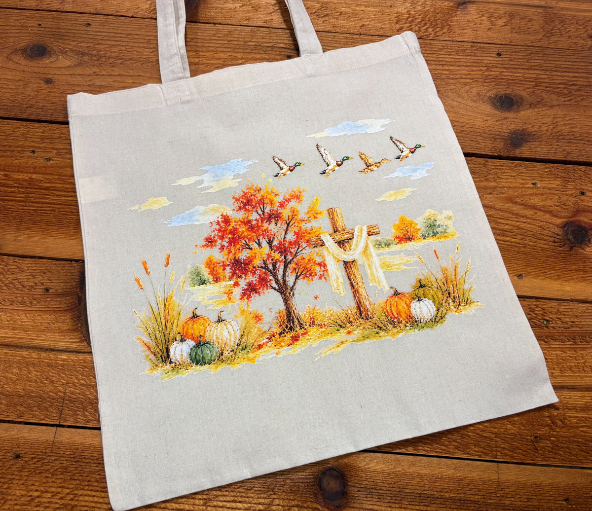 Angled view of a Christian fall tote bag with orange maple tree, draped cross, cattails, and pumpkins on a wood surface
