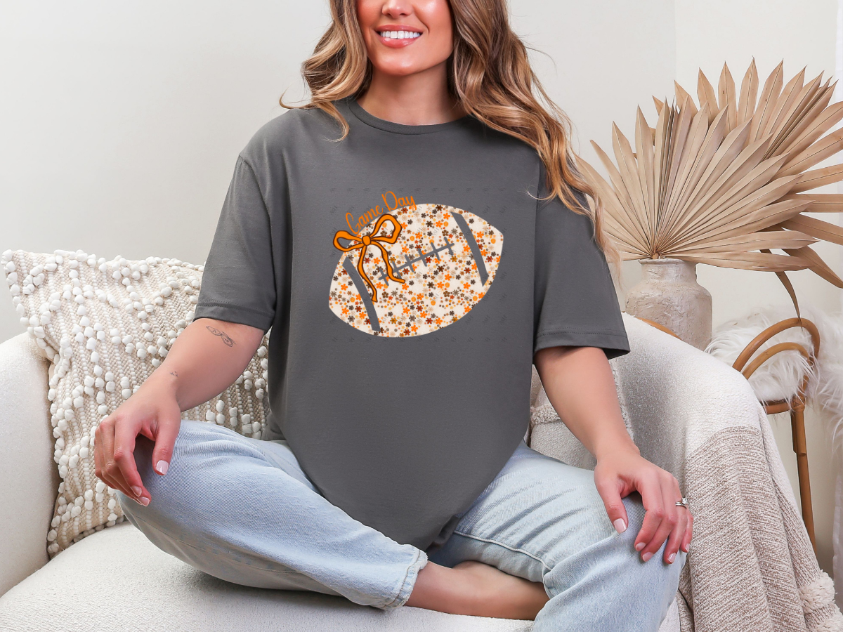 Model wearing a charcoal Floral Football Game Day graphic tee with an orange bow and cream floral football design.