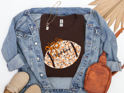 Dark chocolate Floral Football Game Day graphic tee with orange bow, styled with a denim jacket and brown accessories.
