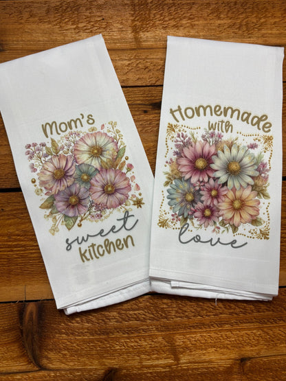 Set of two white floral tea towels reading “Mom’s Sweet Kitchen” and “Homemade with Love” with pastel daisy designs on a wood table