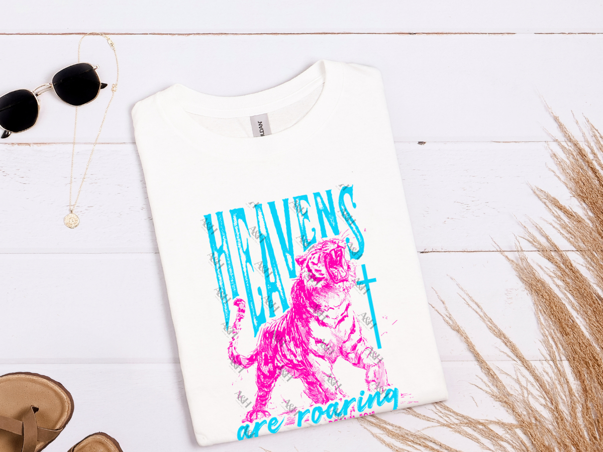 Woman wearing a white Christian graphic tee featuring a roaring tiger, cross, and the phrase "Heavens Are Roaring" with Psalm 19:1 in vibrant turquoise and pink lettering.