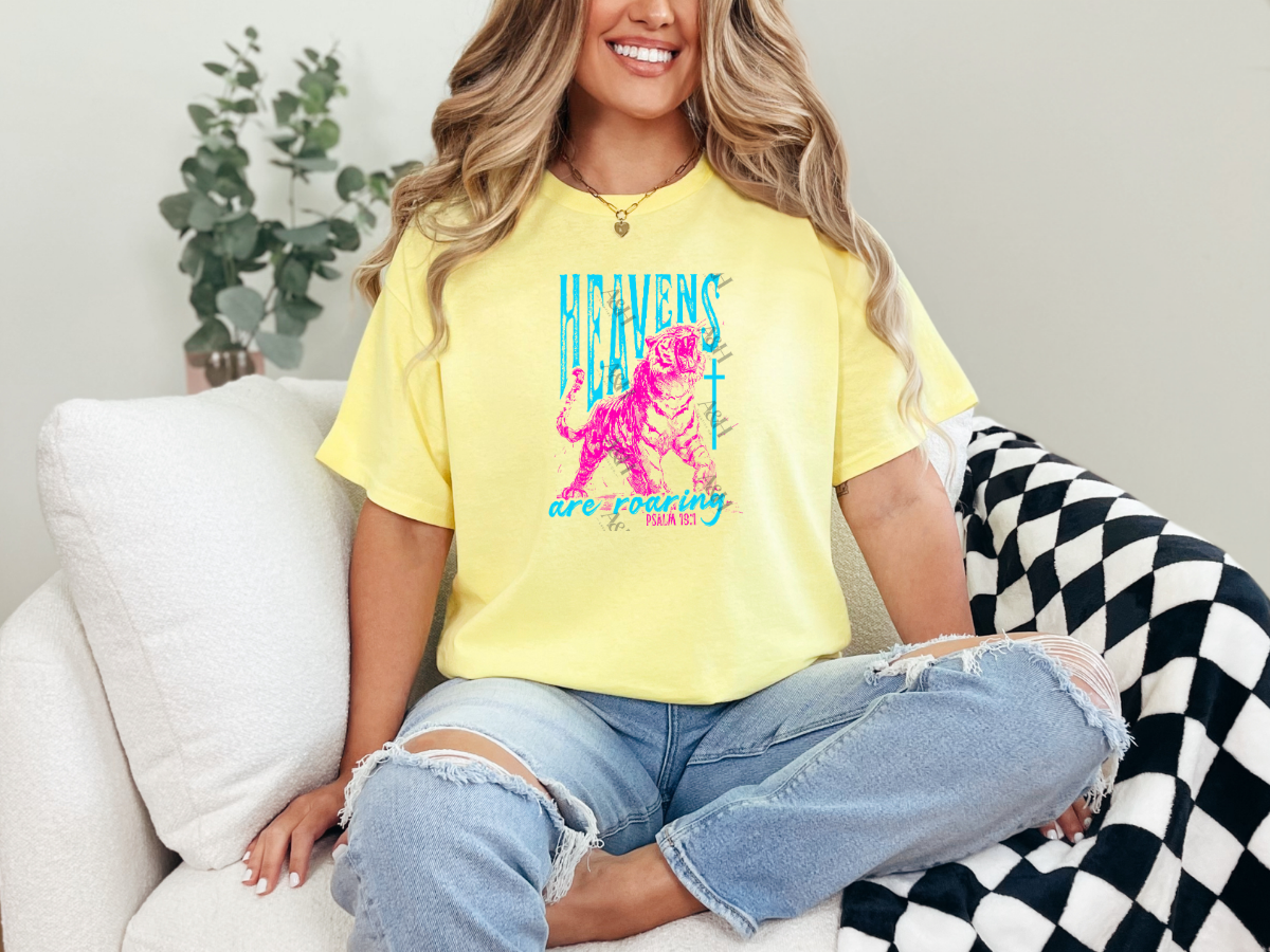 Woman wearing a pastel yellow Christian graphic tee featuring a roaring tiger, cross, and the phrase "Heavens Are Roaring" with Psalm 19:1 in vibrant turquoise and pink lettering.