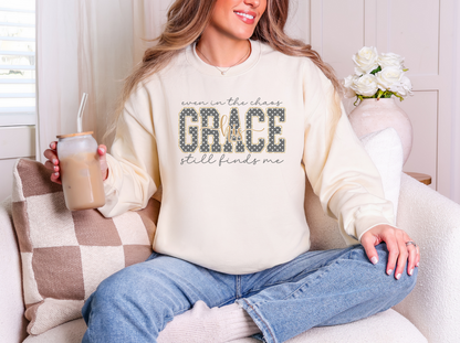 Model wearing a cream crewneck sweatshirt with “Even in the chaos, His grace still finds me” in gray and gold-colored lettering.