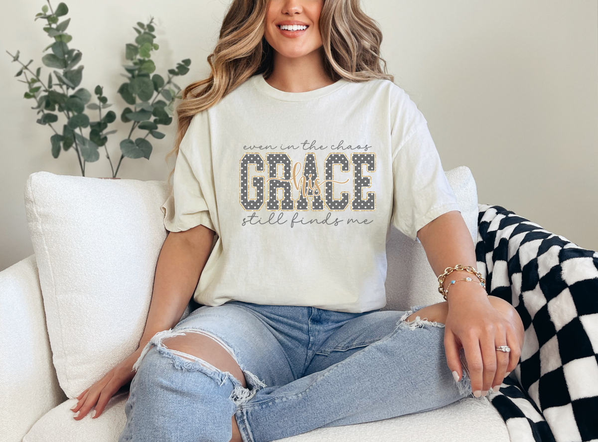 Model wearing a cream Christian T-shirt with “Even in the chaos, His grace still finds me” and polka-dot GRACE lettering.