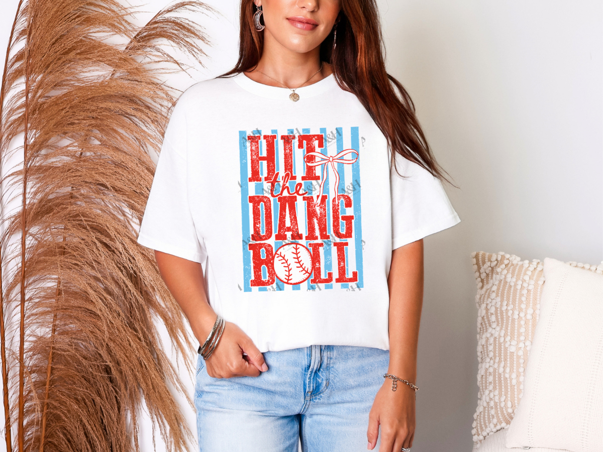 Woman wearing a white Hit the Dang Ball baseball graphic tee with distressed red lettering, blue stripes, baseball, and bow