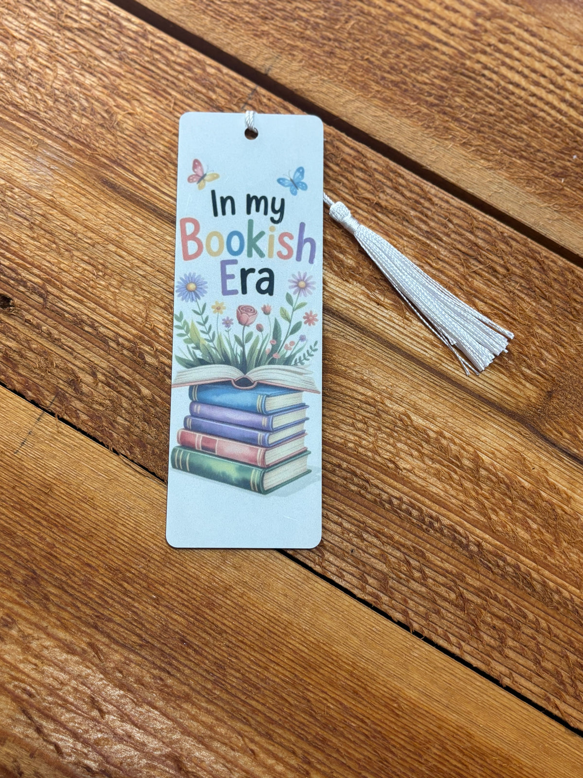 White bookmark reading “In my Bookish Era” with rainbow lettering, floral book stack, butterflies, and white tassel on a wood background