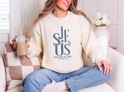 Woman wearing a cream Jesus crewneck sweatshirt with slate blue “Name Above All Names” Philippians 2:9-11 design while holding an iced coffee