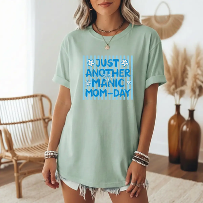 Woman wearing a sage green Manic Mom-Day graphic tee with blue retro lettering and floral accents