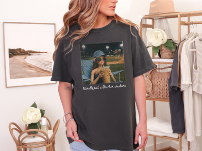 Woman wearing a pepper gray "Literally Just a Bleacher Creature" baseball mom graphic tee featuring an illustrated fan with an oversized iced coffee at a baseball field.
