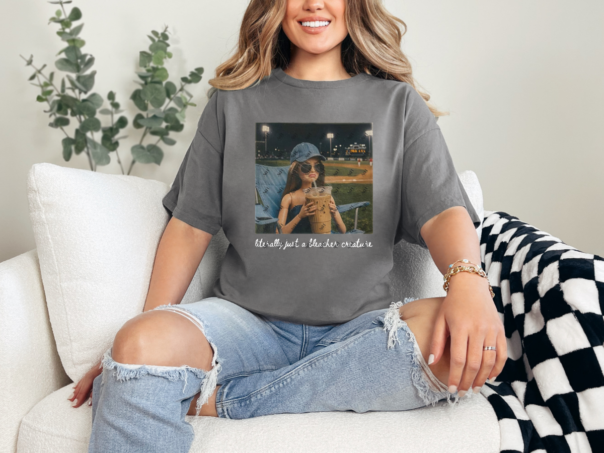 Woman wearing a charcoal gray "Literally Just a Bleacher Creature" baseball mom graphic tee featuring an illustrated fan with an oversized iced coffee at a baseball field.
