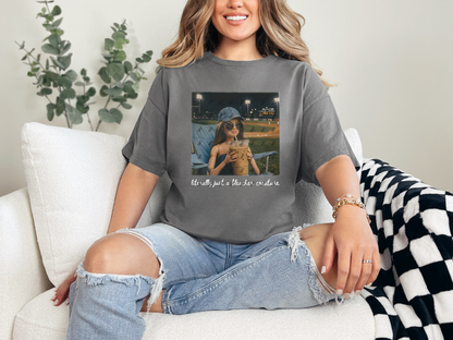Woman wearing a charcoal gray "Literally Just a Bleacher Creature" baseball mom graphic tee featuring an illustrated fan with an oversized iced coffee at a baseball field.
