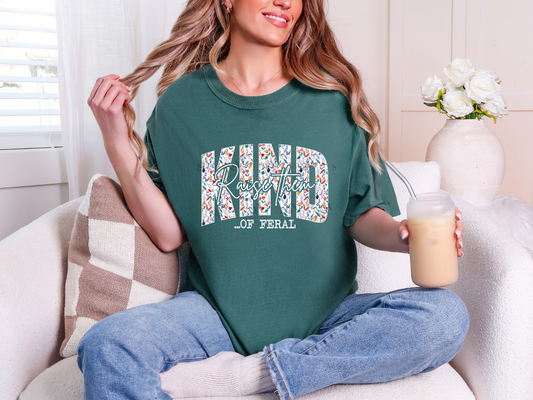 Woman wearing a forest green Raise Them Kind of Feral graphic tee with colorful floral lettering