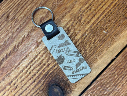 Silver-tone teacher keychain with teach, ABC, inspire, apples, books, pencils, and rulers on a rectangular tag.