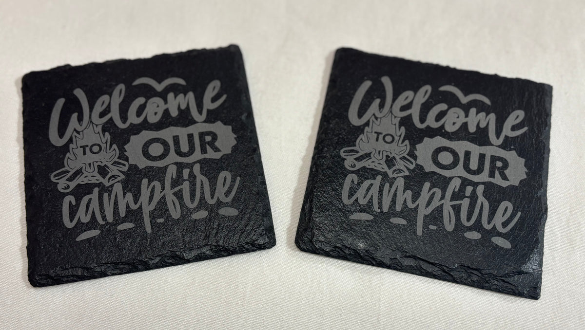 Two square black slate coasters engraved with “Welcome to Our Campfire” lettering and a campfire illustration.